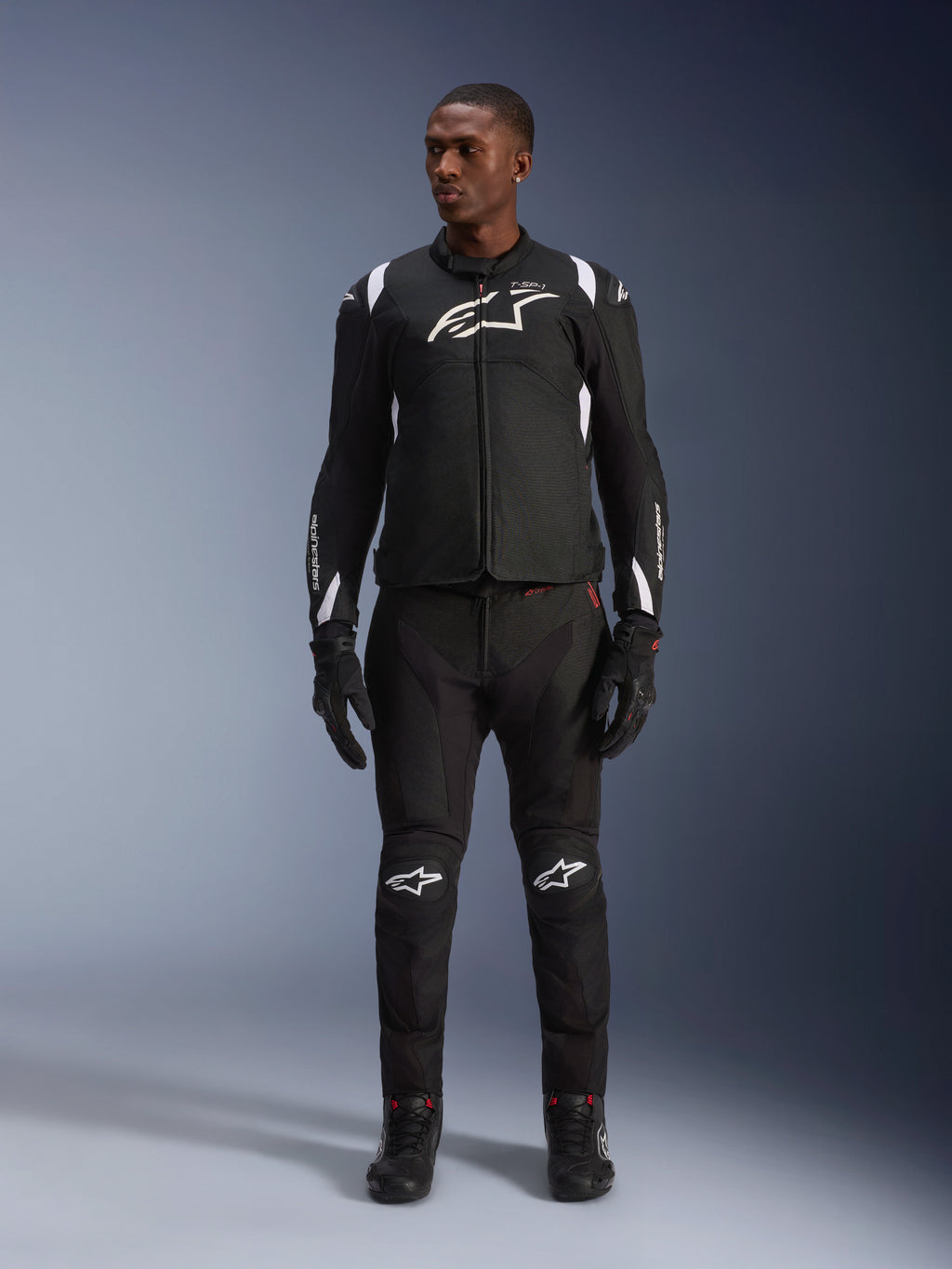 Kurtka Alpinestars T-SP 1 V2 Waterproof, czarno-biała, z wyraźnym białym logo na klatce piersiowej i akcentami na ramionach, noszona z dopasowanymi czarnymi spodniami motocyklowymi i rękawicami dla pełnego zestawu do jazdy szosowej