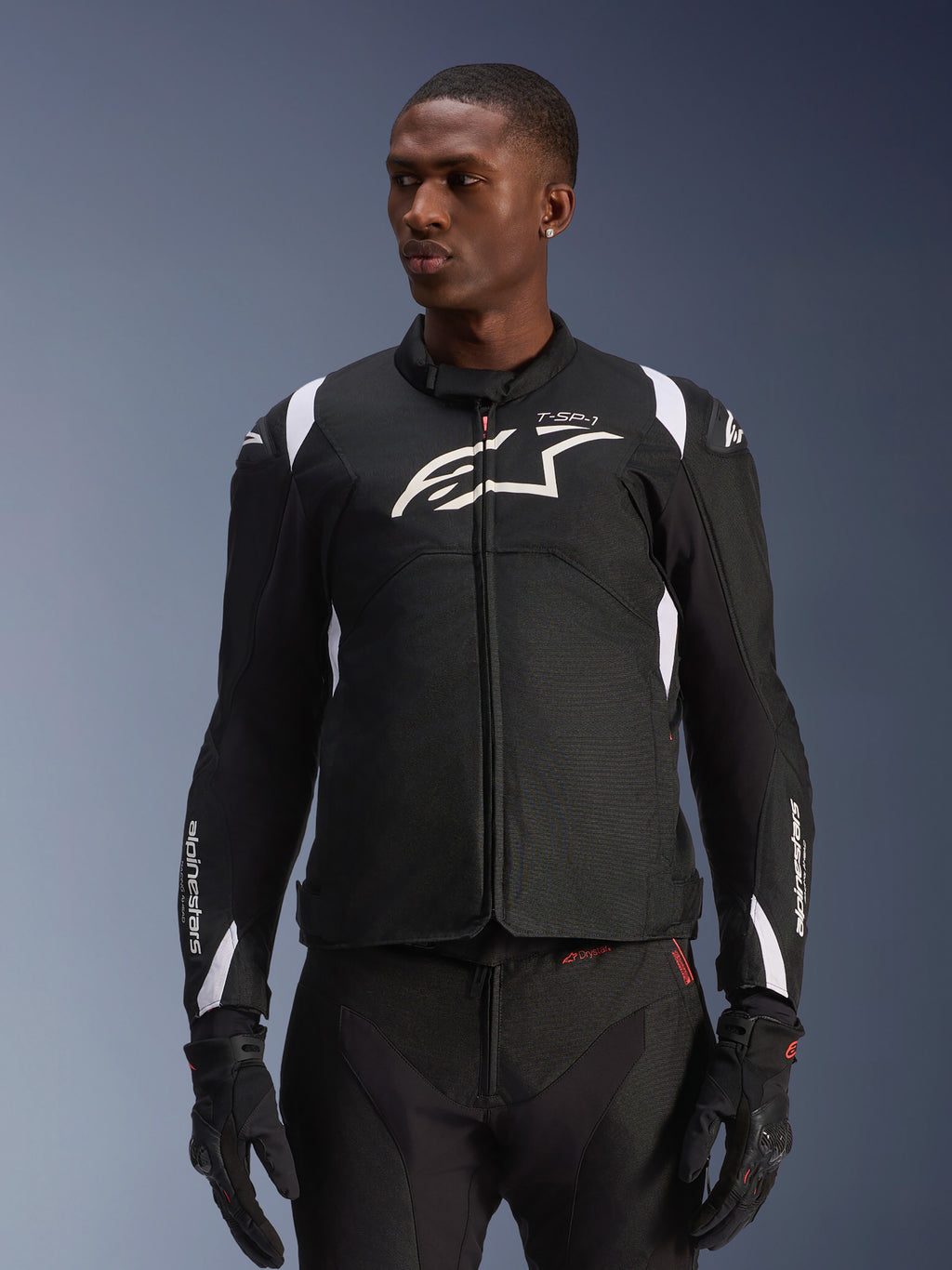 Alpinestars T-SP 1 V2 Waterproof Jacket, sportowa tekstylna kurtka motocyklowa, czarno-biała z subtelnymi czerwonymi detalami, noszona przez modela z dopasowanymi rękawicami i spodniami, białe logo Alpinestars na piersi, branding T-SP-1, ochronna sylwetka do jazdy szosowej