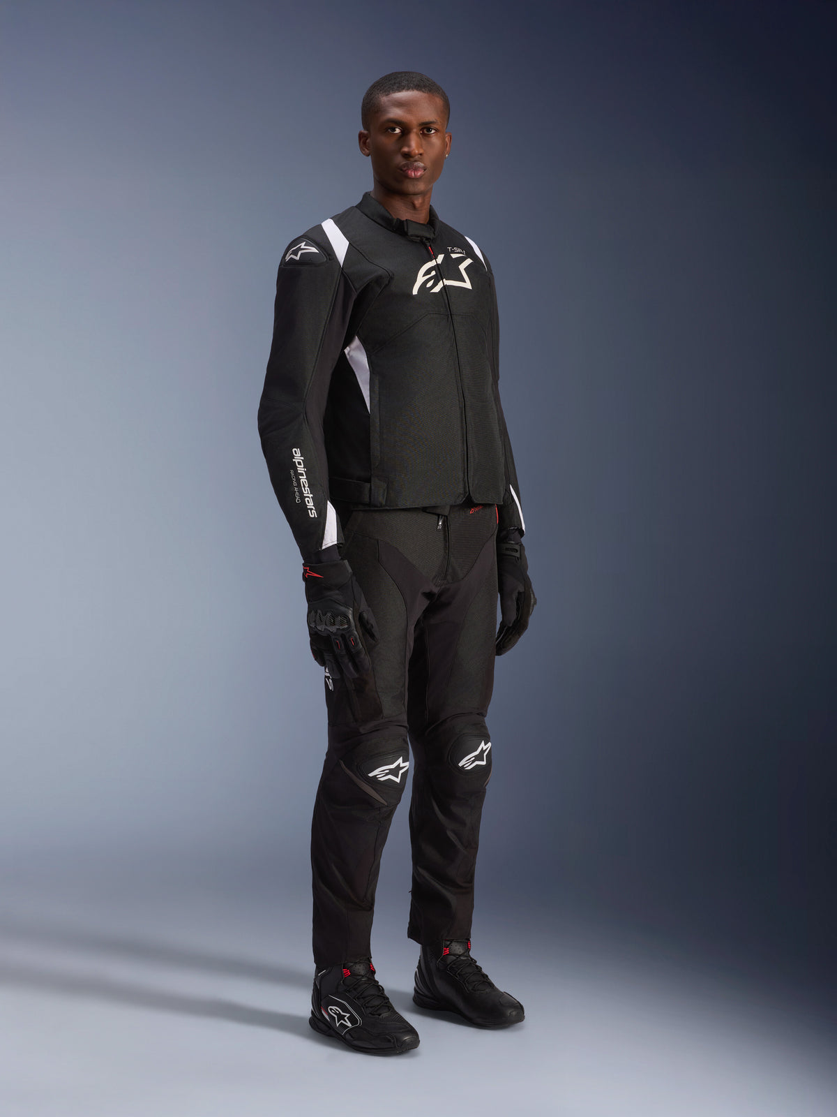 Alpinestars T-SP 1 V2 Waterproof Jacket, czarno-biała, męski model w kompletnym stroju z czarnymi spodniami motocyklowymi, rękawicami i butami, zdjęcie studyjne całej sylwetki, sportowa kurtka tekstylna z białymi logotypami i dopasowanym krojem 3D.