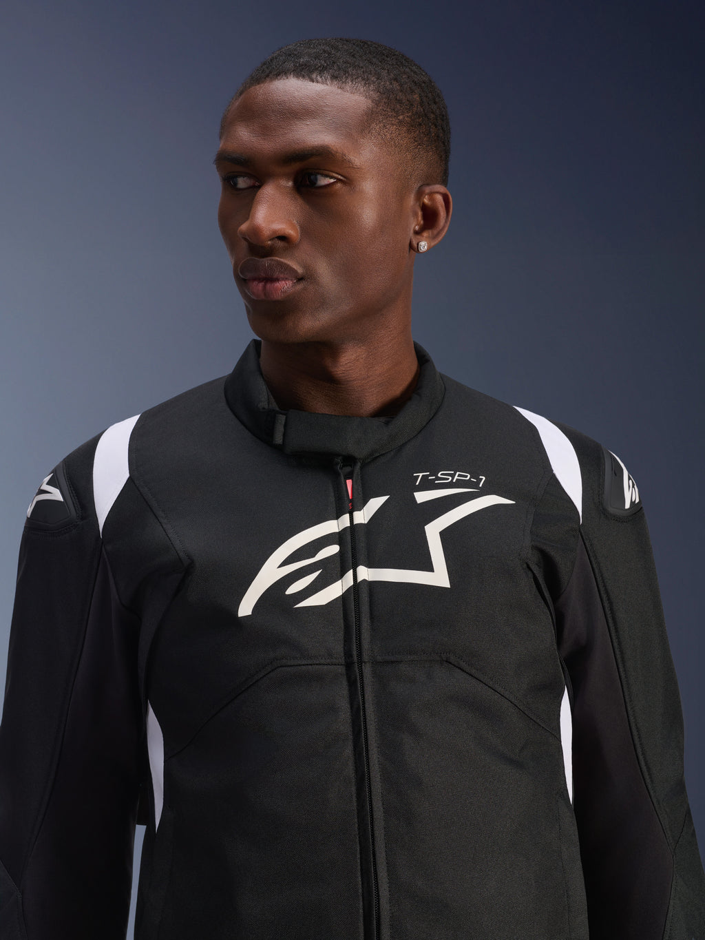Kurtka Alpinestars T-SP 1 V2 Waterproof prezentowana przez modela, czarno-biała tekstylna kurtka motocyklowa, duże białe logo Alpinestars i branding T-SP-1 na klatce piersiowej, sportowy design z ochronnymi ślizgaczami na ramionach