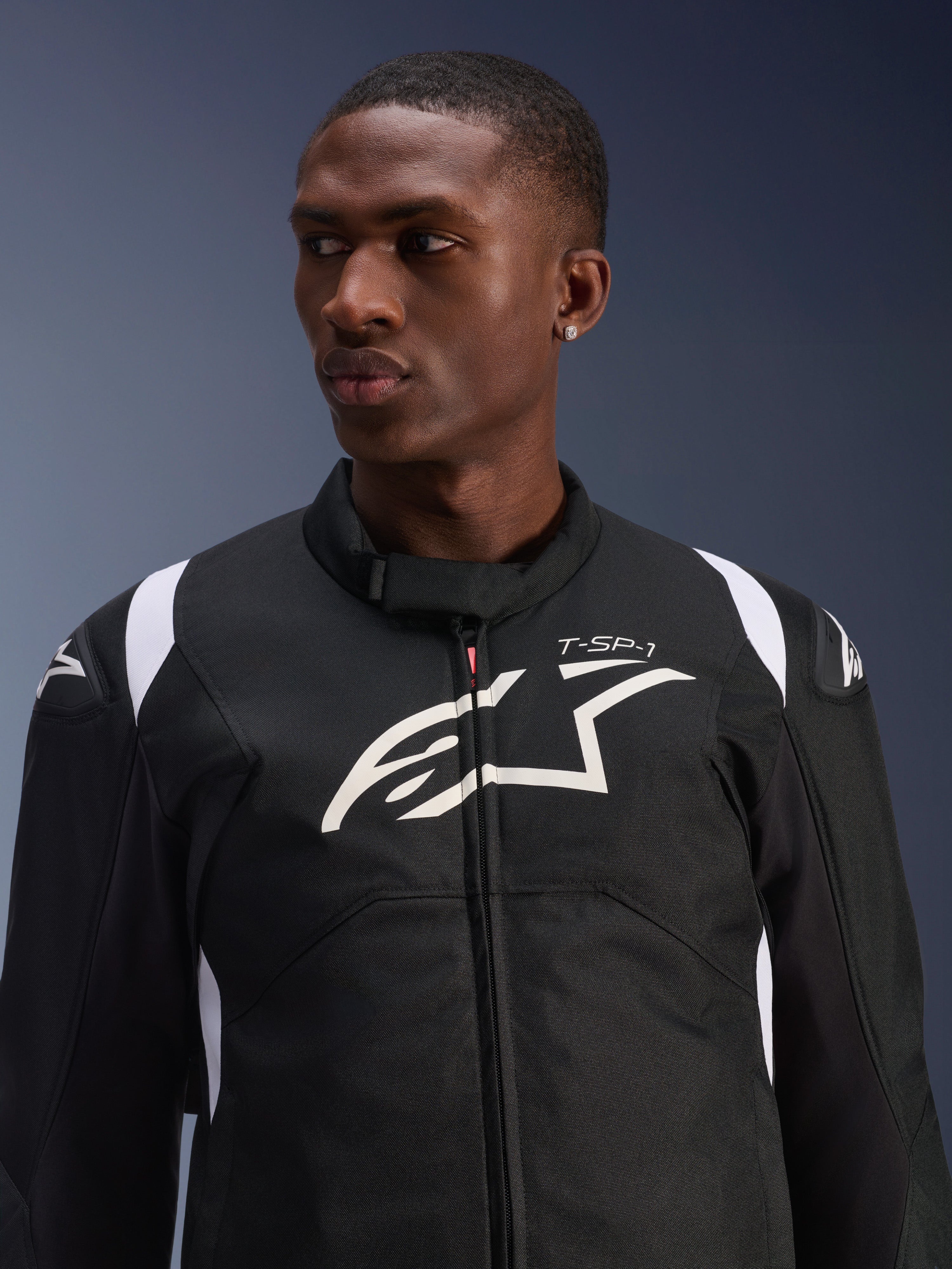 Kurtka Alpinestars T-SP 1 V2 Waterproof prezentowana przez modela, czarno-biała tekstylna kurtka motocyklowa, duże białe logo Alpinestars i branding T-SP-1 na klatce piersiowej, sportowy design z ochronnymi ślizgaczami na ramionach
