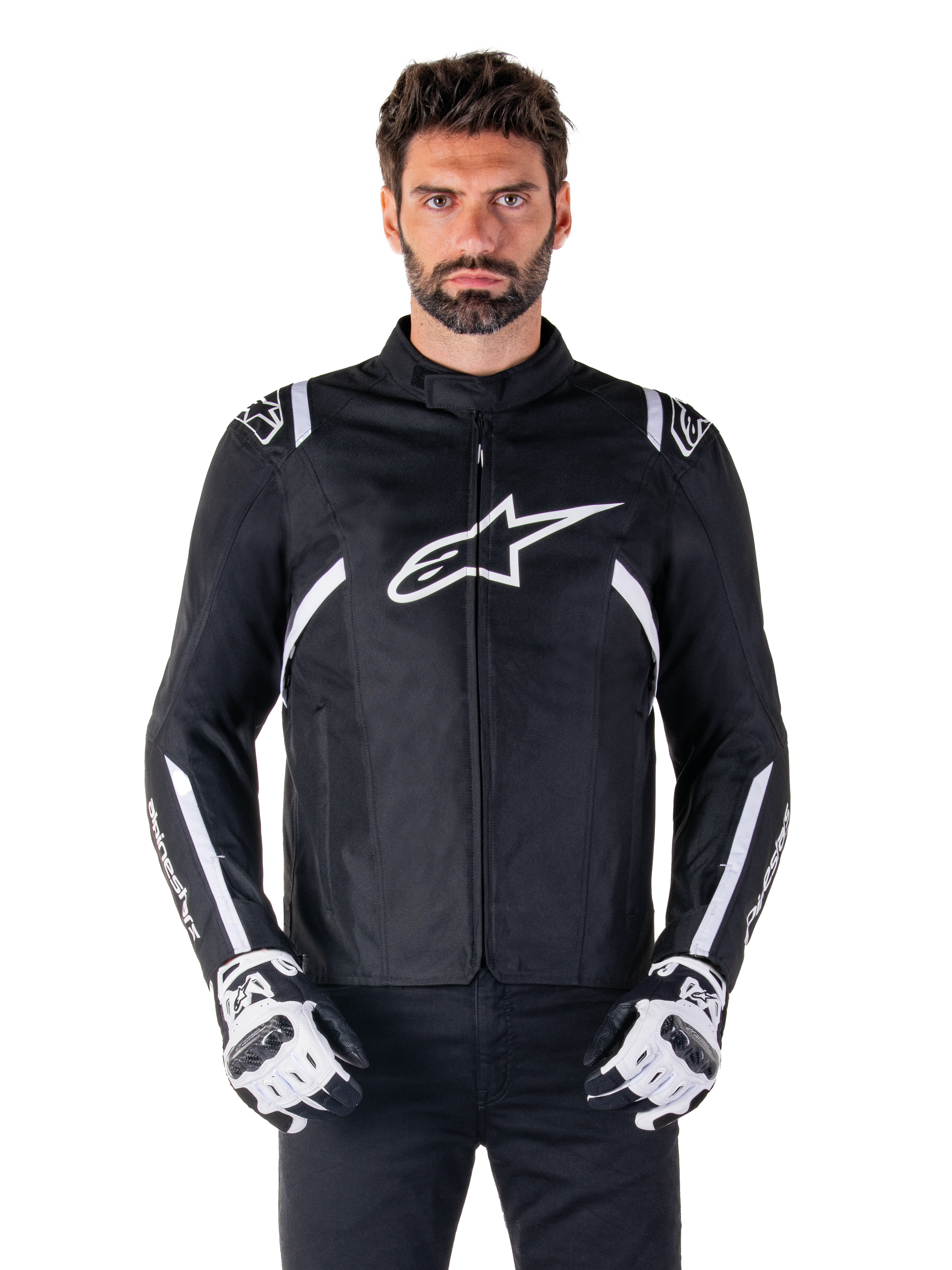 Alpinestars T-SPS V2 Waterproof Jacket w kolorze czarno-białym, widok z przodu na modelu, wodoodporna tekstylna kurtka motocyklowa z białymi pasami na ramionach i rękawach, duże białe logo Alpinestars na piersi, noszona z ochronnymi rękawicami motocyklowymi, smukła sylwetka inspirowana sportem