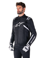 Kurtka Alpinestars T-SPS V2 Waterproof, czarno-biała, noszona przez modela w pasujących rękawicach motocyklowych, tekstylna kurtka o sportowym kroju z białym logo Alpinestars na piersi i ramionach, branding "alpinestars T-SPS" na rękawie, stylowa odzież motocyklowa inspirowana wyścigami