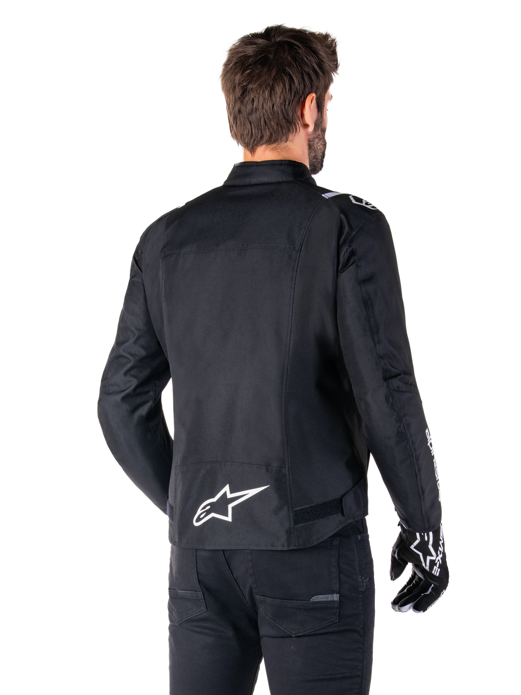 Alpinestars T-SPS V2 Waterproof Jacket, czarno-biała wodoodporna kurtka motocyklowa, widok z tyłu na motocyklistę w kurtce, białe logo Alpinestars na dole pleców, tekstylna konstrukcja o sportowym kroju, zestawiona z czarnymi spodniami i rękawicami