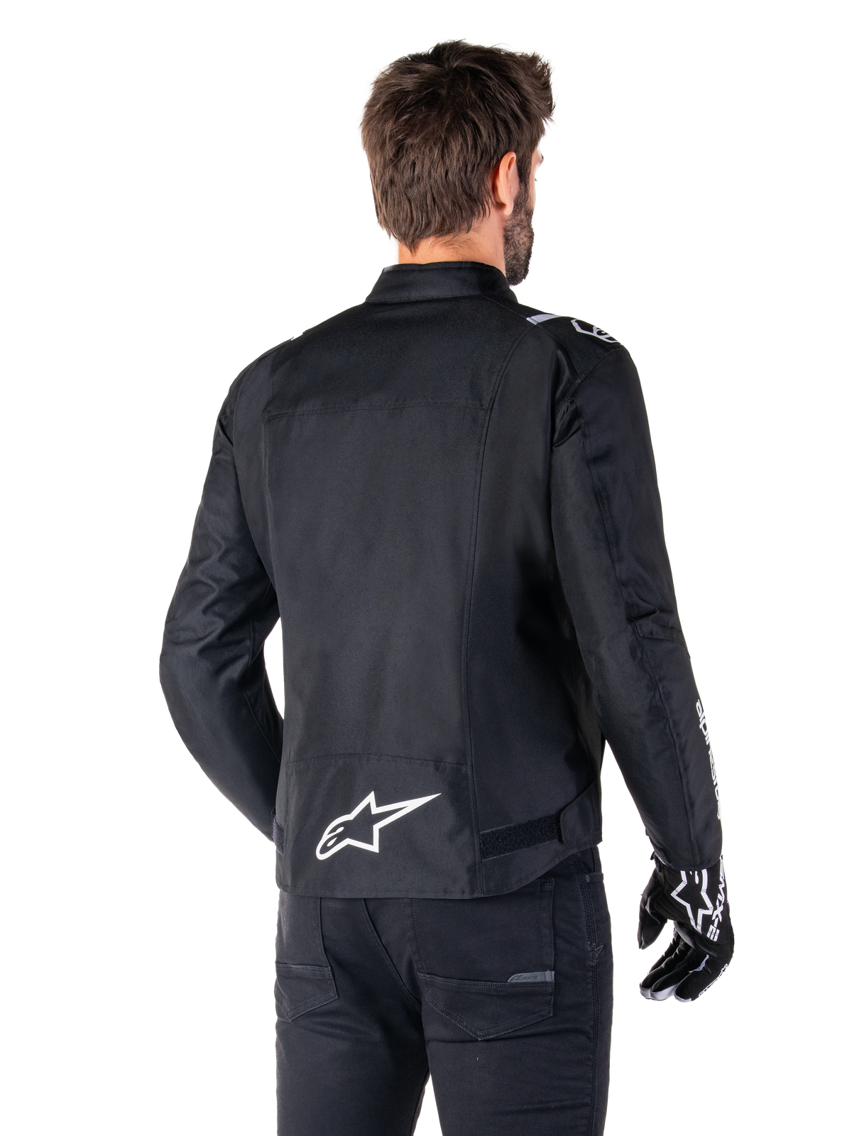 Alpinestars T-SPS V2 Waterproof Jacket, czarno-biała wodoodporna kurtka motocyklowa, widok z tyłu na motocyklistę w kurtce, białe logo Alpinestars na dole pleców, tekstylna konstrukcja o sportowym kroju, zestawiona z czarnymi spodniami i rękawicami