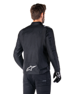 Alpinestars T-SPS V2 Waterproof Jacket, czarno-biała wodoodporna kurtka motocyklowa, widok z tyłu na motocyklistę w kurtce, białe logo Alpinestars na dole pleców, tekstylna konstrukcja o sportowym kroju, zestawiona z czarnymi spodniami i rękawicami