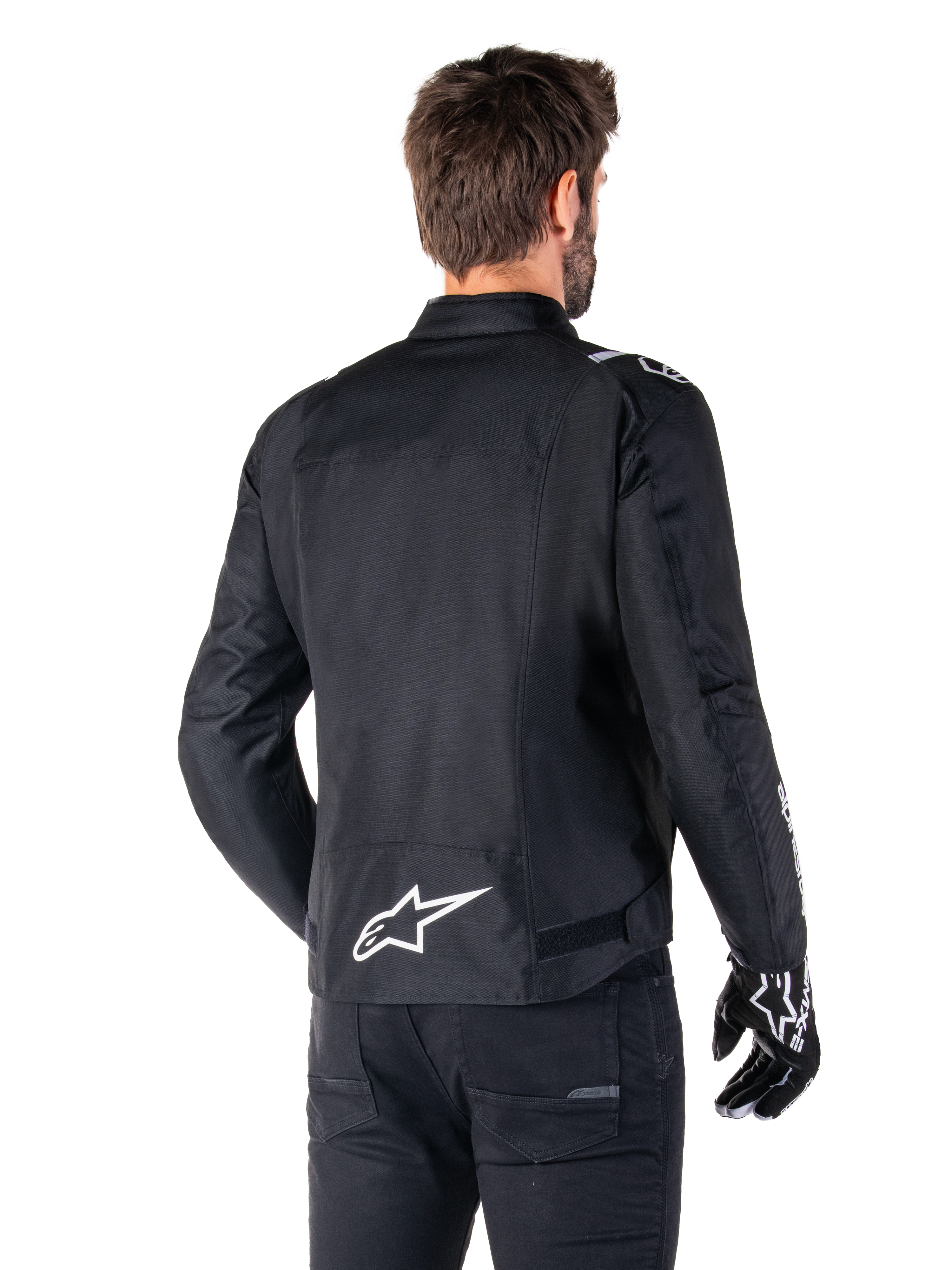 Alpinestars T-SPS V2 Waterproof Jacket, czarno-biała wodoodporna kurtka motocyklowa, widok z tyłu na motocyklistę w kurtce, białe logo Alpinestars na dole pleców, tekstylna konstrukcja o sportowym kroju, zestawiona z czarnymi spodniami i rękawicami