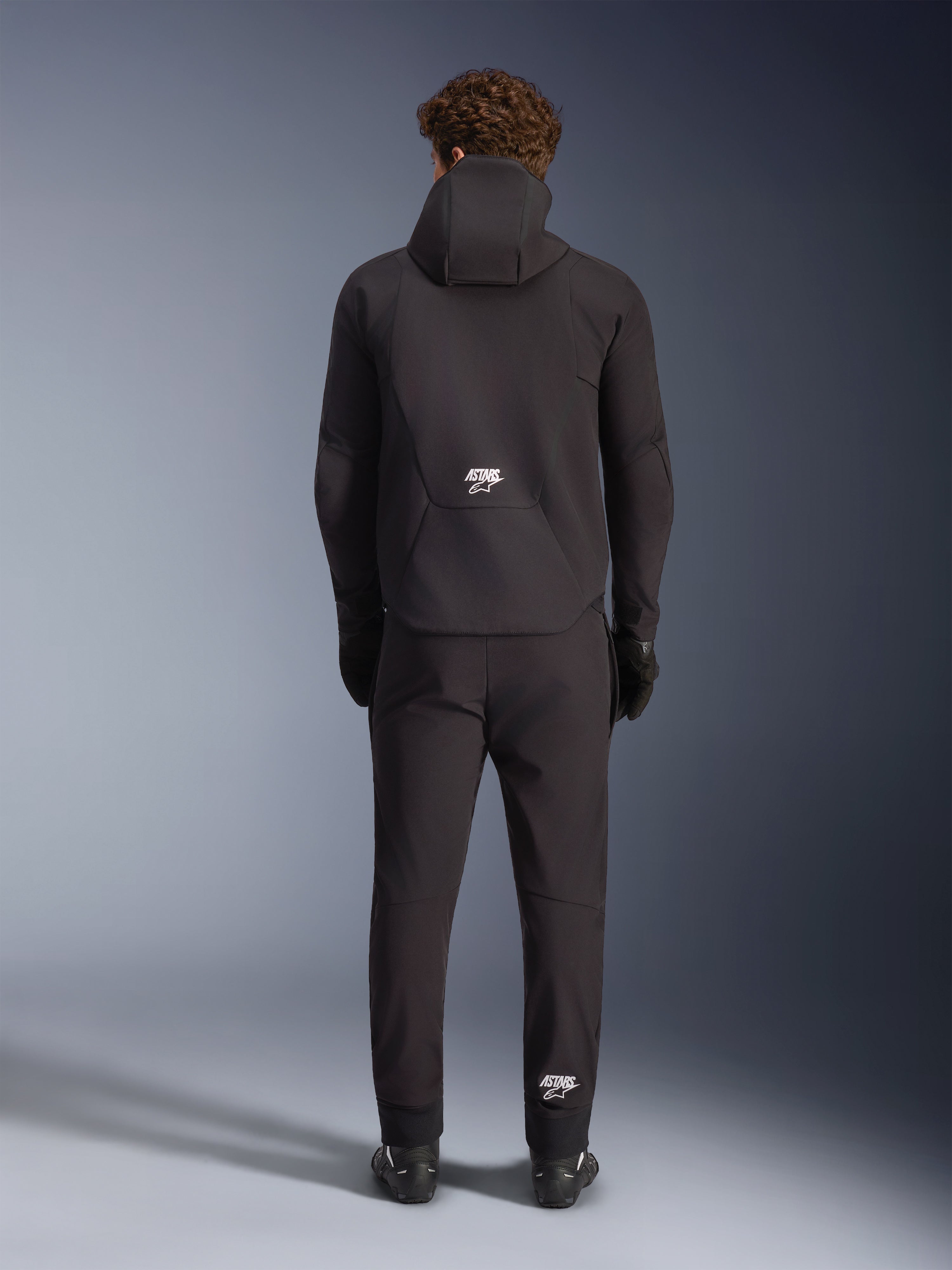 Alpinestars Aeroshell Waterproof Jacket, czarna, widziana od tyłu na modelu noszącym dopasowane czarne spodnie i rękawice motocyklowe, sportowa konstrukcja typu softshell z kapturem i białymi logo ASTARS, nowoczesna odzież motocyklowa