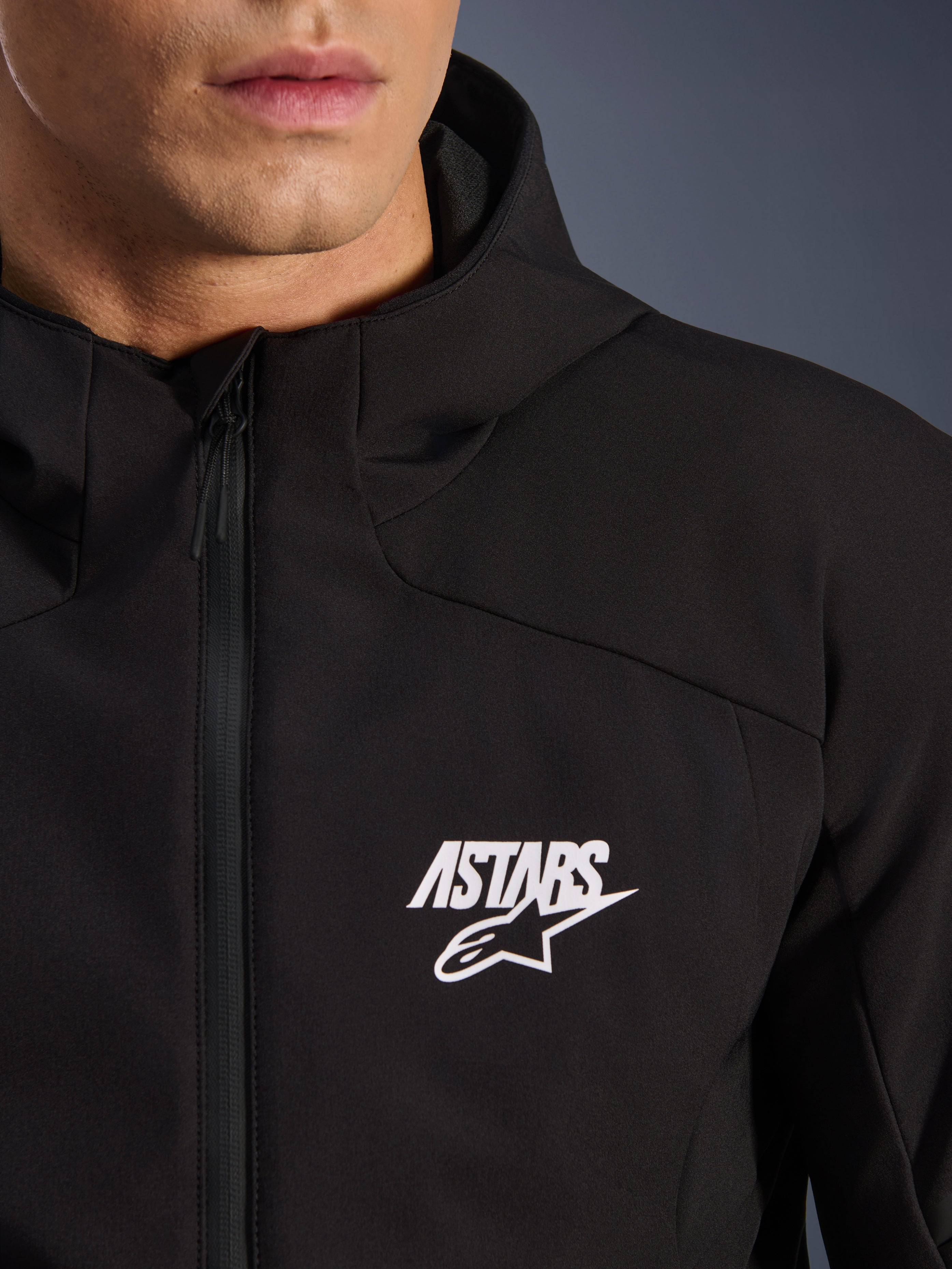 Alpinestars Aeroshell Waterproof Jacket, czarna motocyklowa kurtka typu softshell, zbliżenie na klatkę piersiową z białym logo gwiazdy ASTARS i wodoodpornym przednim zamkiem błyskawicznym, zintegrowany kaptur, nowoczesny sportowy design