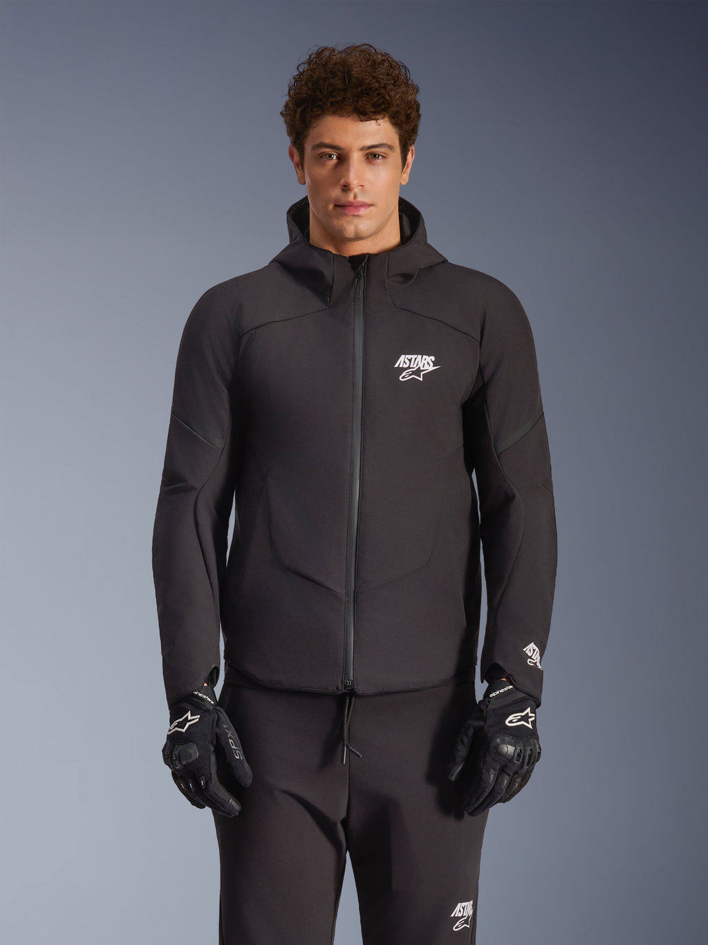 Alpinestars Aeroshell Waterproof Jacket, czarna miejska kurtka motocyklowa, fason z kapturem i pełnym zamkiem z przodu, białe logotypy na piersi i rękawach, zestawiona z dopasowanymi czarnymi spodniami i rękawicami