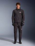 Męski model w czarnej kurtce Alpinestars Aeroshell Waterproof, czarne spodnie techniczne i rękawice motocyklowe, fason softshell z kapturem i białymi akcentami logo, nowoczesny strój inspirowany sportami motorowymi do jazdy szosowej