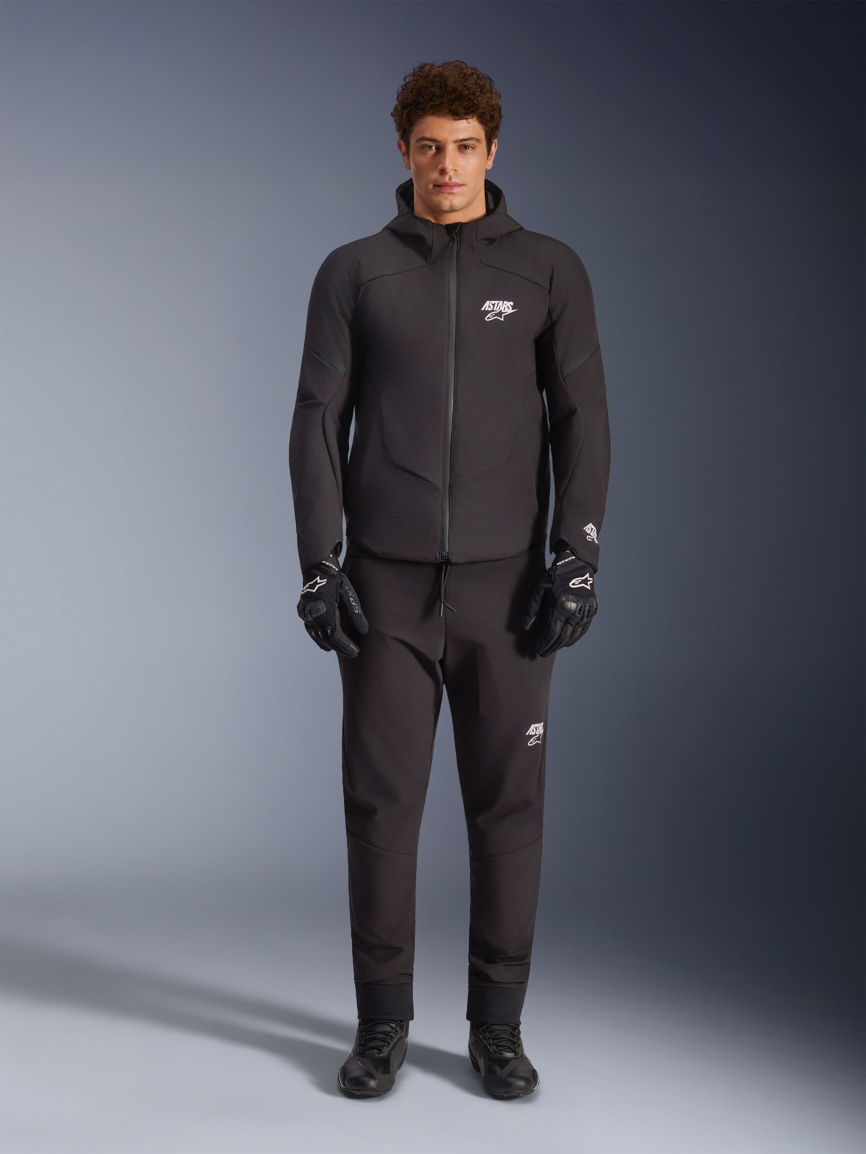 Męski model w czarnej kurtce Alpinestars Aeroshell Waterproof, czarne spodnie techniczne i rękawice motocyklowe, fason softshell z kapturem i białymi akcentami logo, nowoczesny strój inspirowany sportami motorowymi do jazdy szosowej