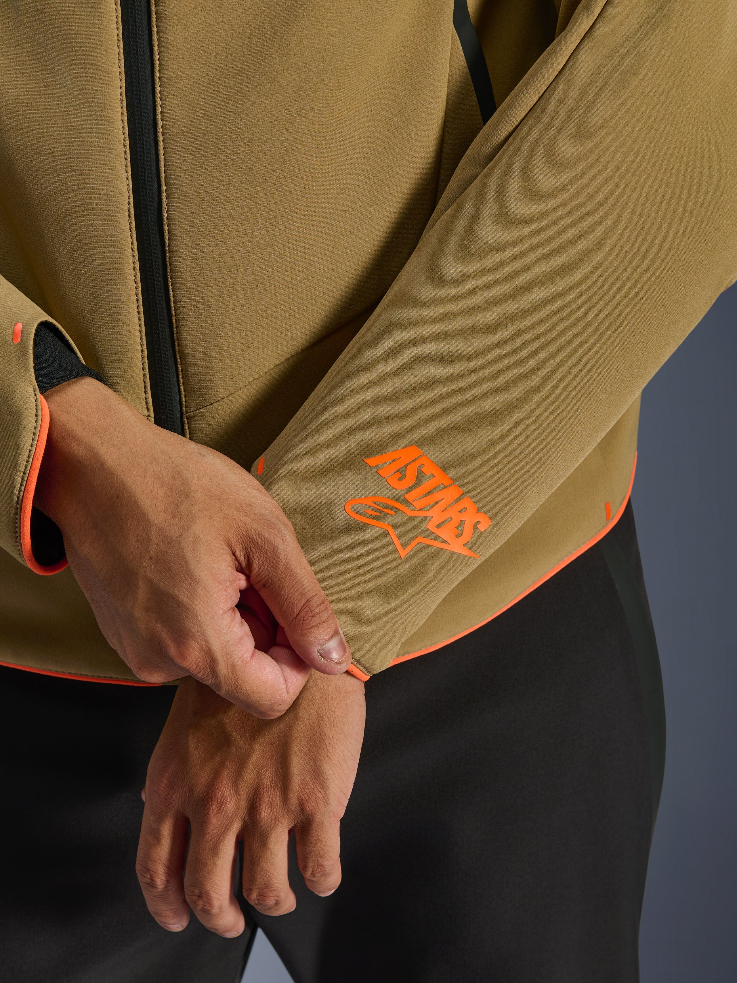 Alpinestars Aeroshell Waterproof Jacket, kolor ciemny khaki z pomarańczowymi akcentami fluo, zbliżenie na mankiet rękawa i dolną krawędź z jaskrawopomarańczowym logo Astars, neonową lamówką i wodoodpornym zamkiem błyskawicznym, sportowa konstrukcja softshell.
