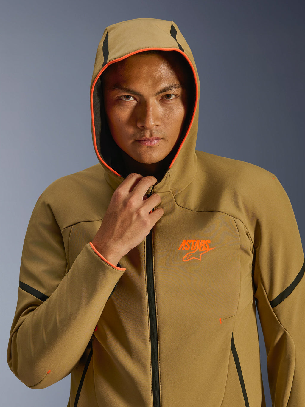 Kurtka Alpinestars Aeroshell Waterproof, Dark Khaki Black Orange Fluo, model mający na sobie kurtkę z założonym kapturem, jaskrawe neonowe pomarańczowe wykończenie i pomarańczowe logo ASTARS, czarne akcenty, nowoczesny wodoodporny softshell w sportowym stylu