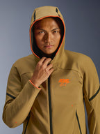 Kurtka Alpinestars Aeroshell Waterproof, Dark Khaki Black Orange Fluo, model mający na sobie kurtkę z założonym kapturem, jaskrawe neonowe pomarańczowe wykończenie i pomarańczowe logo ASTARS, czarne akcenty, nowoczesny wodoodporny softshell w sportowym stylu
