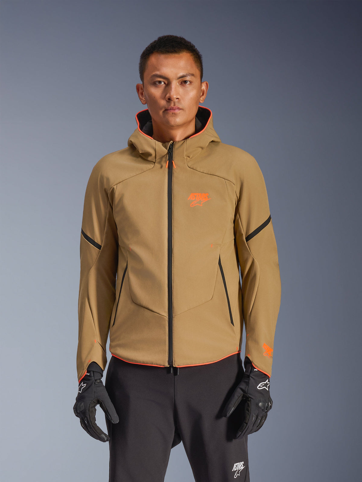 Alpinestars Aeroshell Waterproof Jacket, kurtka motocyklowa z kapturem w kolorze ciemnego khaki z pomarańczowymi logotypami i czarnymi zamkami, czarne linie akcentowe na rękawach, pokazana z czarnymi rękawicami i spodniami