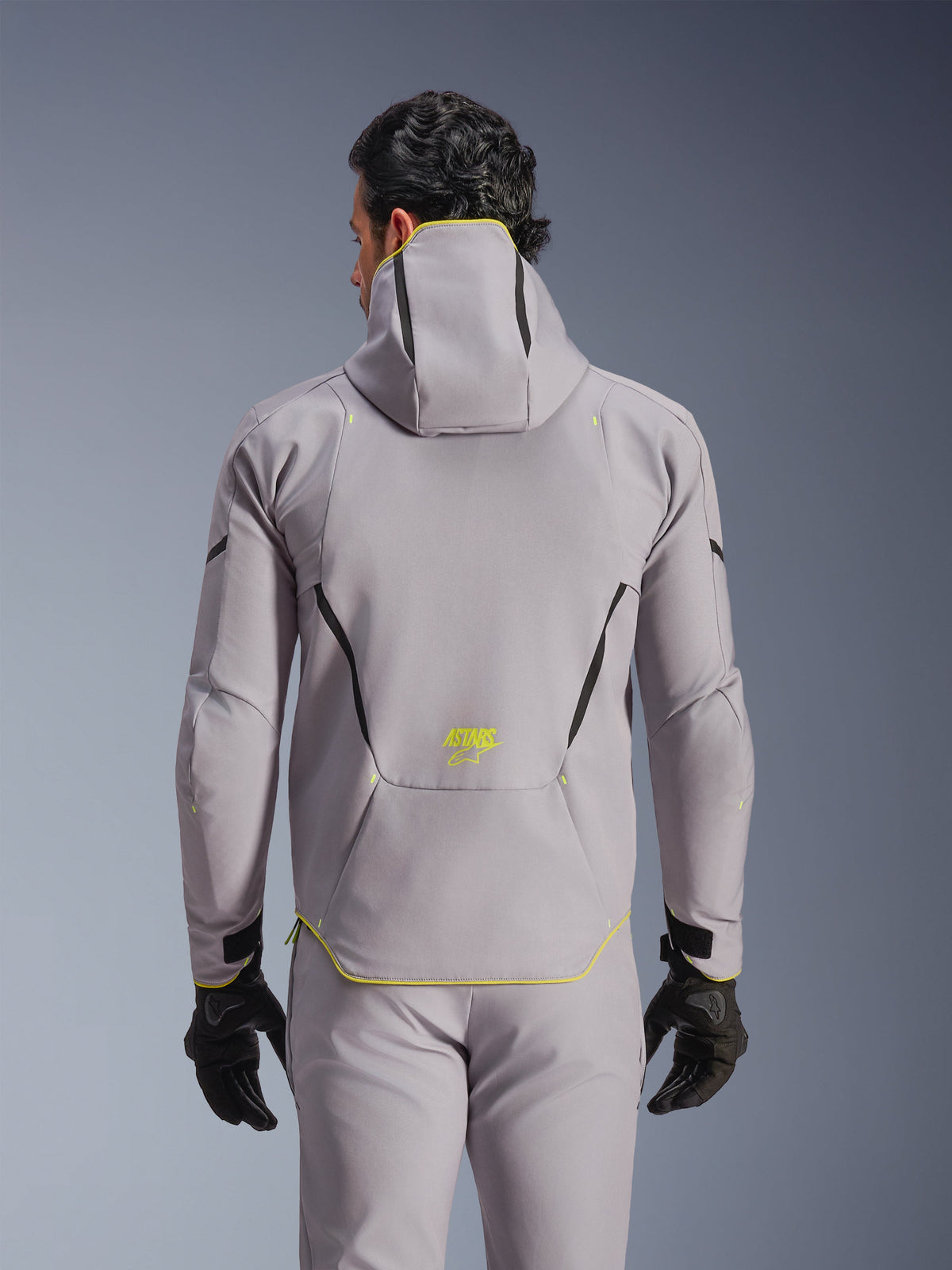 Alpinestars Aeroshell Waterproof Jacket, wodoodporna motocyklowa kurtka softshell, średnioszara z neonowożółtymi akcentami, widok z tyłu pokazujący zintegrowany kaptur i czarne kontrastowe linie, żółte logo ASTARS na dole pleców, noszona z pasującymi szarymi spodniami i czarnymi rękawicami