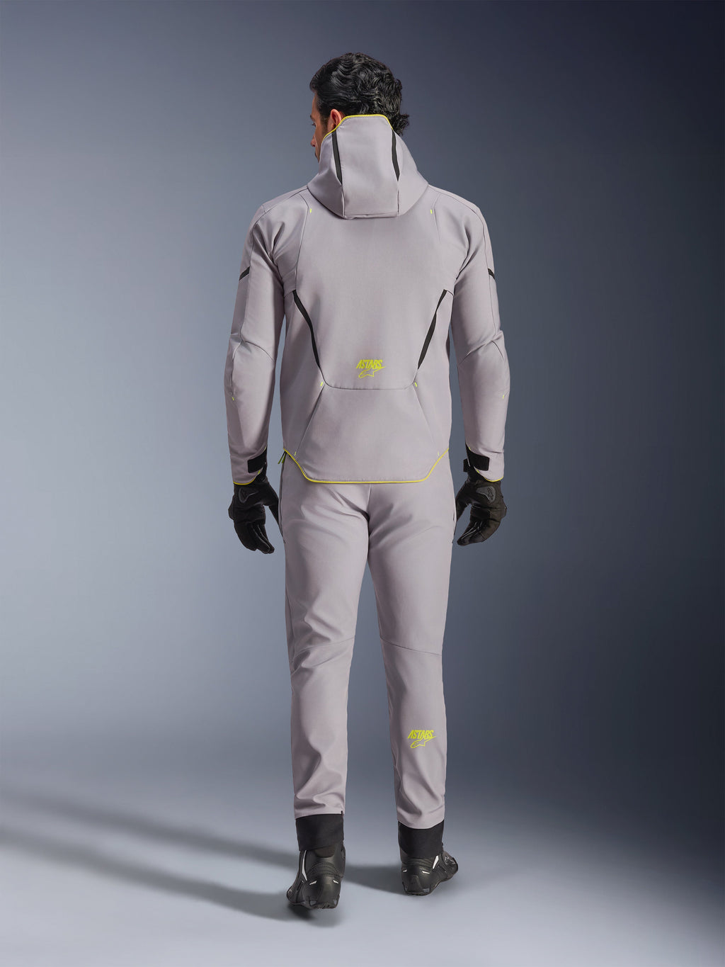 Wodoodporna kurtka Alpinestars Aeroshell w kolorze Mid Gray Yellow Fluo, pełny widok z tyłu, noszona z pasującymi szarymi spodniami, czarnymi rękawicami i czarnymi butami motocyklowymi. Posiada konstrukcję softshell z kapturem, jaskrawożółte akcenty oraz logo 'ASTARS', zaprojektowana z myślą o nowoczesnych osiągach motocyklowych na drodze.