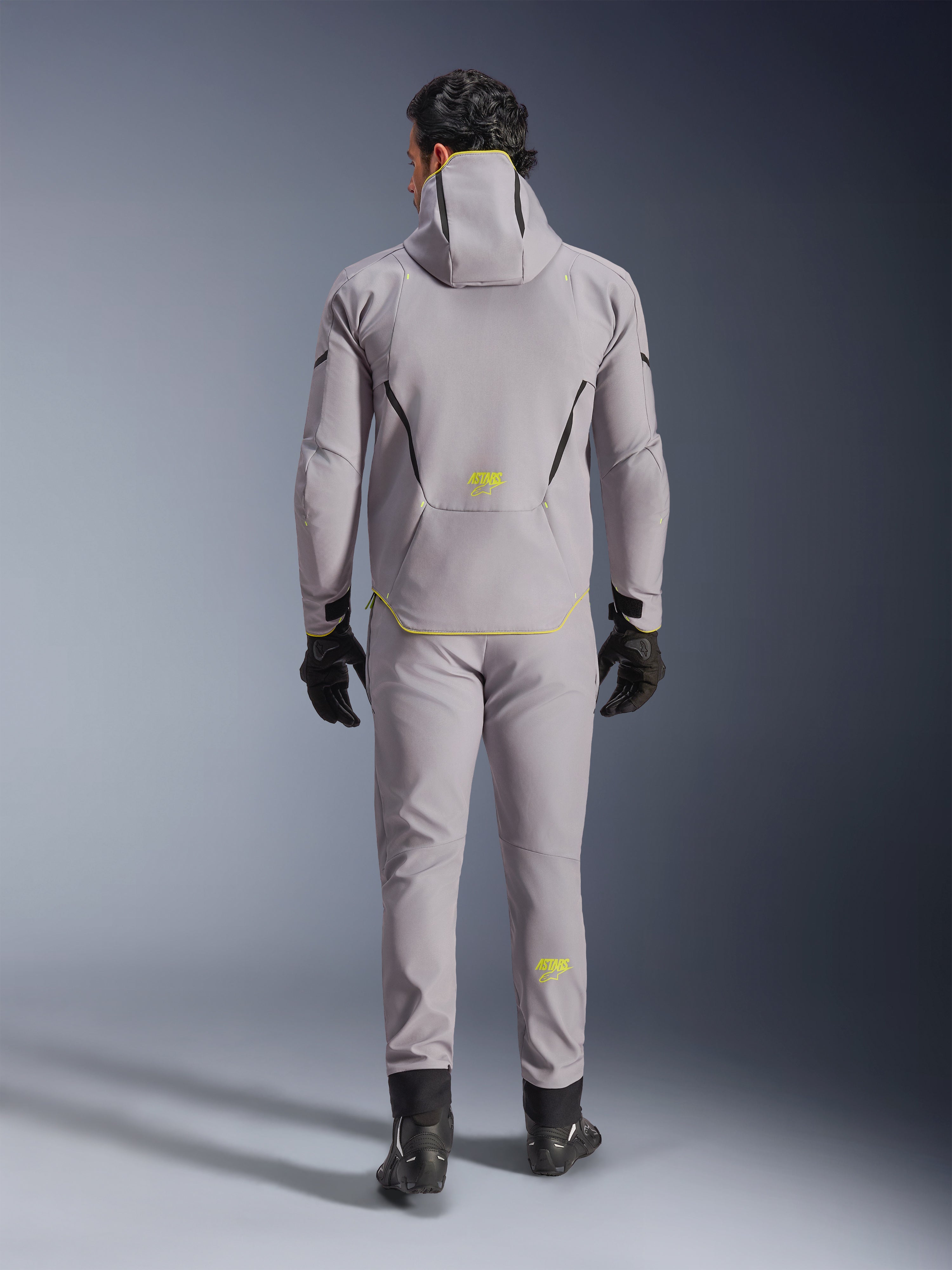 Wodoodporna kurtka Alpinestars Aeroshell w kolorze Mid Gray Yellow Fluo, pełny widok z tyłu, noszona z pasującymi szarymi spodniami, czarnymi rękawicami i czarnymi butami motocyklowymi. Posiada konstrukcję softshell z kapturem, jaskrawożółte akcenty oraz logo 'ASTARS', zaprojektowana z myślą o nowoczesnych osiągach motocyklowych na drodze.