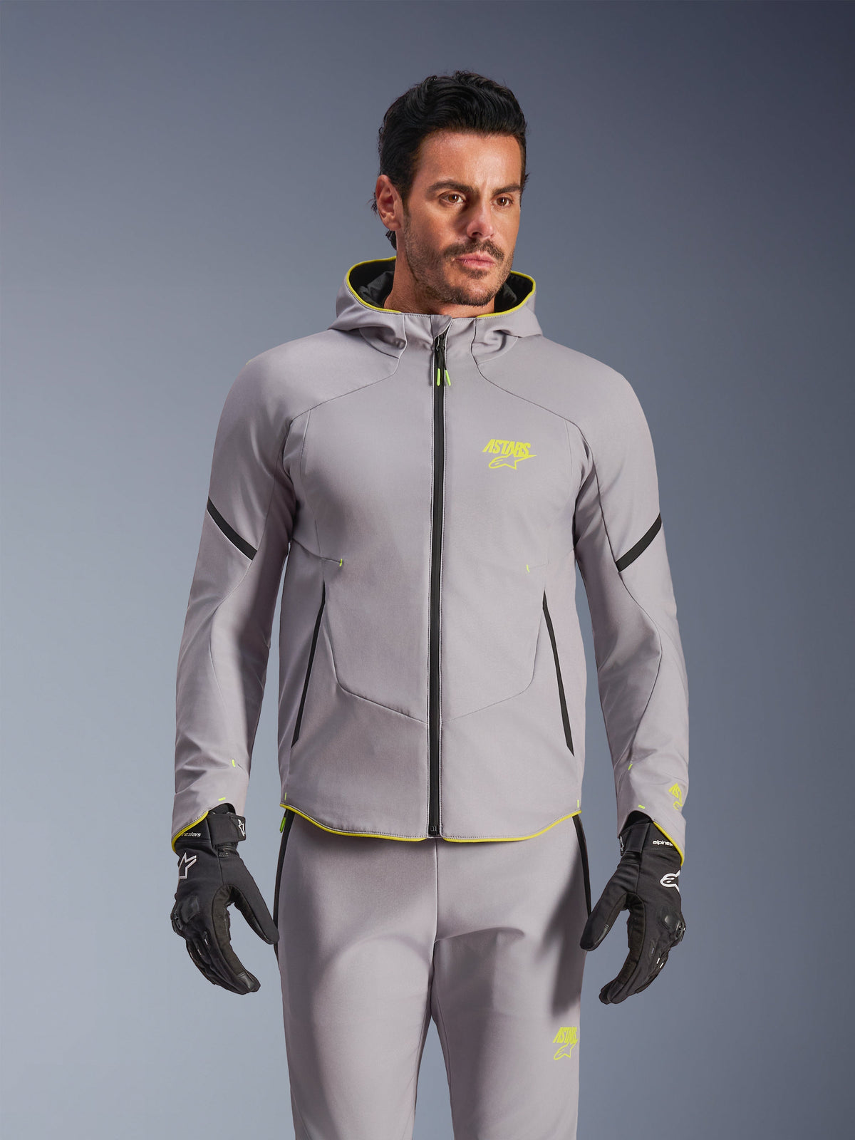 Alpinestars Aeroshell Waterproof Jacket, średnioszara i żółta neonowa, motocyklowa kurtka softshell z kapturem i czarnymi zamkami, noszona z pasującymi szarymi spodniami i czarnymi rękawicami, wodoodporna odzież do jazdy szosowej