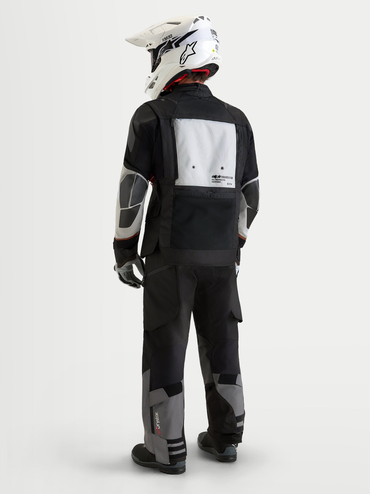 Alpinestars Harok V2 Drystar, męska kurtka motocyklowa adventure, ciemnoszara, jasnoszara i czarna, widok z tyłu przedstawiający modułowy panel wentylacyjny, wzmocnienia PU oraz pasujące spodnie adventure i biały kask off-road