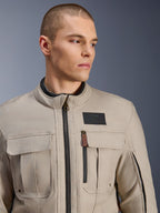 Męski model w kurtce Alpinestars Borrego Drystar® w kolorze Warm Gray, motocyklowa kurtka adventure o klasycznym wyglądzie woskowanego płótna, z funkcjonalnymi kieszeniami cargo na klatce piersiowej i czarną naszywką z logo Alpinestars.