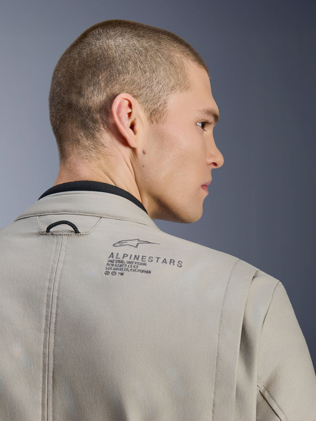 Kurtka Alpinestars Borrego Drystar® w kolorze Warm Gray, zbliżenie na tył ramienia, teksturowany materiał z woskowanego płótna, czarne logo Alpinestars i napis heritage, prezentowana przez modela, kurtka motocyklowa adventure