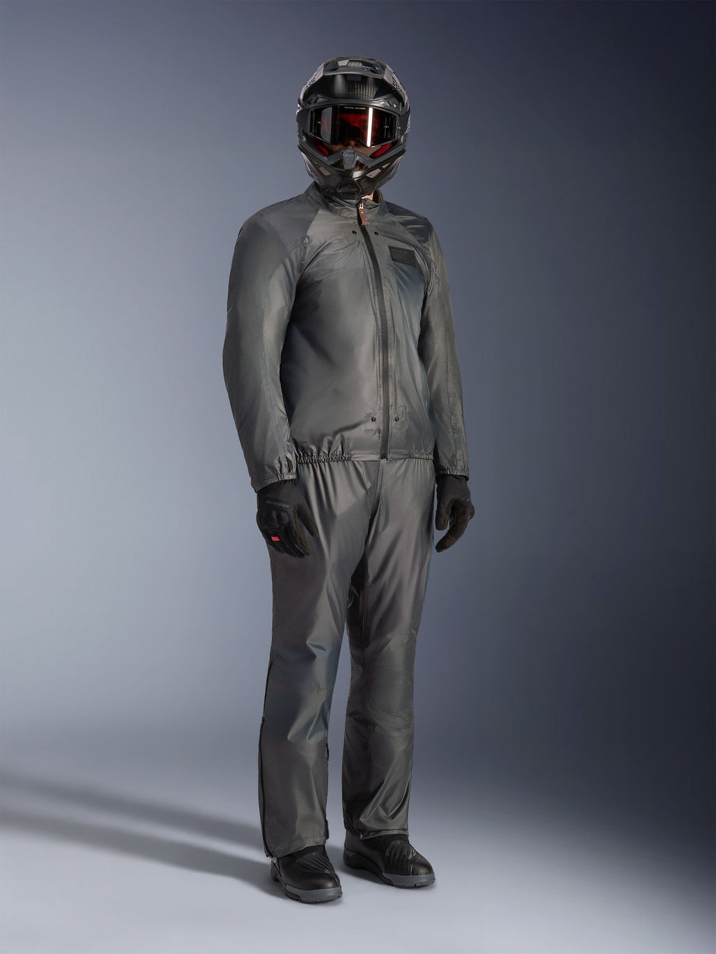 Motocyklista w kurtce Alpinestars Borrego Drystar® w kolorze Warm Gray, kompletny strój adventure z dopasowanymi szarymi spodniami, czarny kask z goglami i czarne rękawice, elegancki wodoodporny design tekstylny
