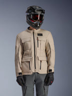 Alpinestars Borrego Drystar® Jacket, turystyczna kurtka motocyklowa Adventure, kolor Warm Gray (ciepły szary), z wieloma przednimi kieszeniami cargo i czarnymi zamkami błyskawicznymi, noszona z czarnym kaskiem, goglami i ciemnoszarymi spodniami motocyklowymi, wytrzymały tekstylny design turystyczny