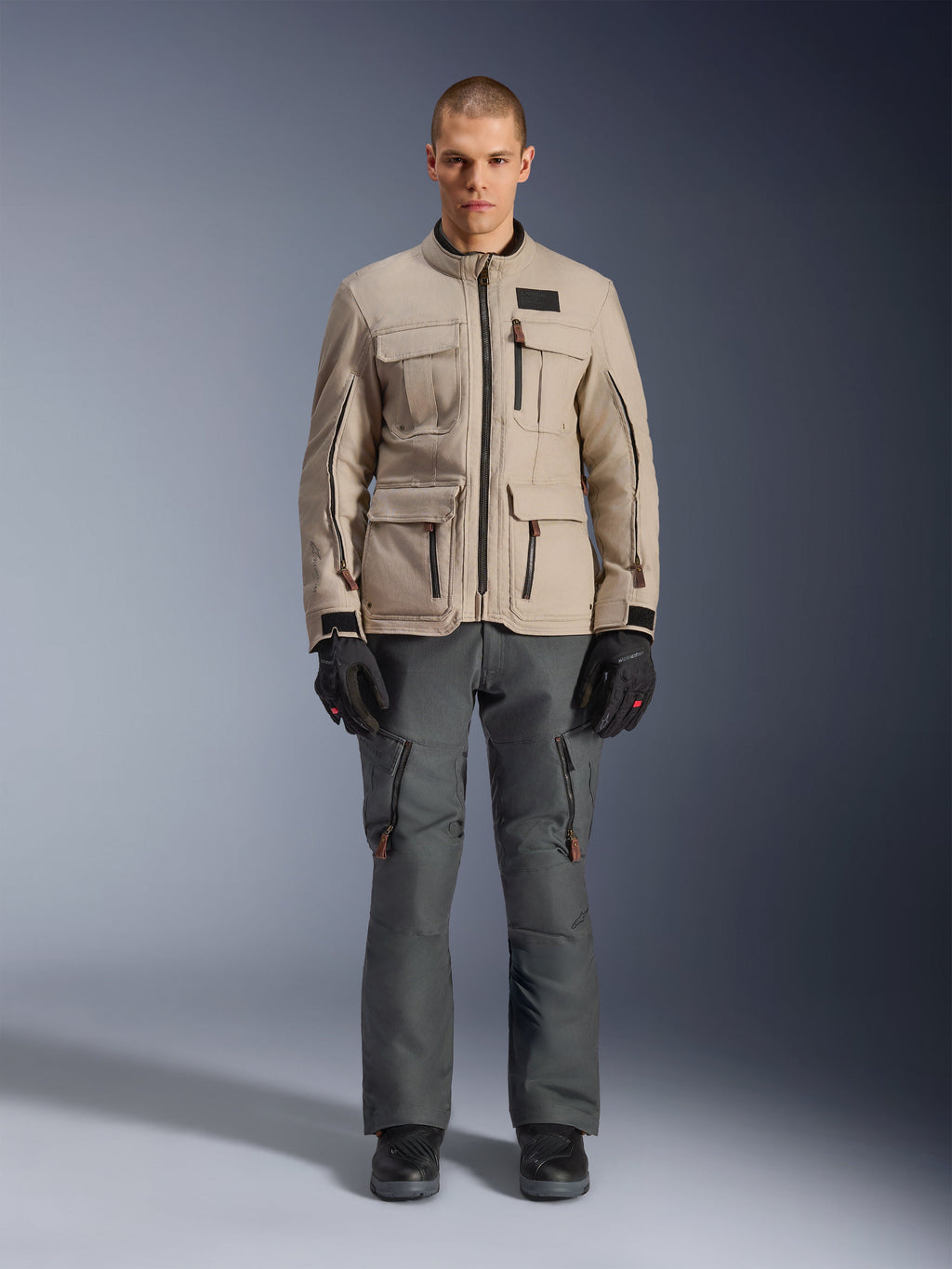 Kurtka Alpinestars Borrego Drystar®, motocyklowa kurtka adventure, kolor Warm Gray, noszona przez modela z ciemnoszarymi spodniami motocyklowymi i czarnymi rękawicami, wiele kieszeni cargo, trwała konstrukcja tekstylna o klasycznym wyglądzie woskowanego płótna