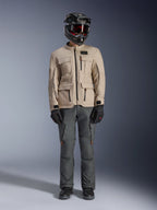 Kurtka Alpinestars Borrego Drystar® w kolorze Warm Gray, przygodowa kurtka motocyklowa z czterema przednimi kieszeniami cargo, zestawiona z ciemnoszarymi spodniami motocyklowymi, czarnym kaskiem off-road i czarnymi rękawicami, o wyglądzie wytrzymałego woskowanego płótna