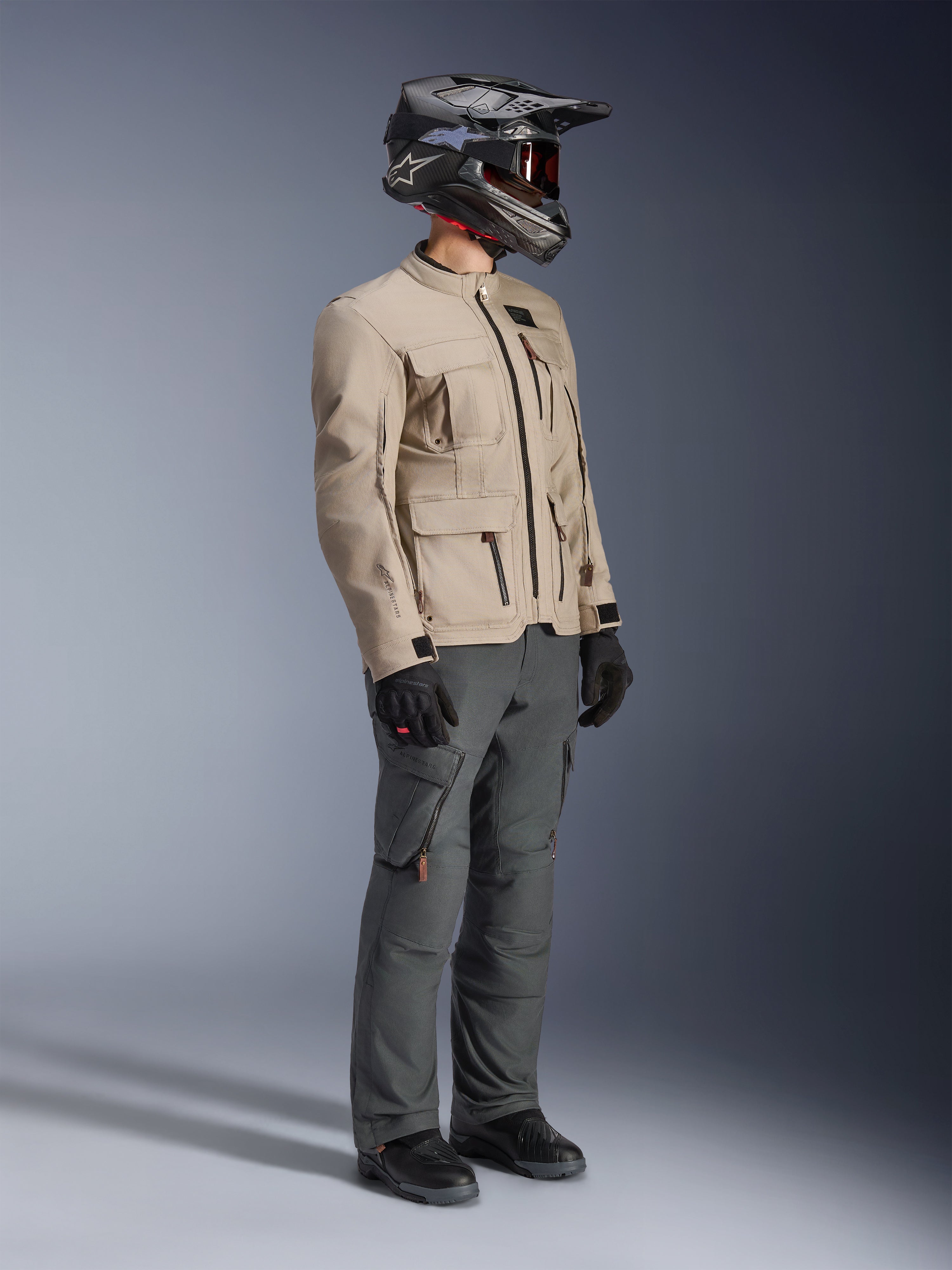 Motocyklista w kurtce Alpinestars Borrego Drystar® w kolorze Warm Gray, turystyczna kurtka motocyklowa o teksturze woskowanego płótna z kieszeniami użytkowymi, zestawiona z ciemnoszarymi spodniami motocyklowymi, kaskiem z włókna węglowego, czarnymi rękawicami i czarnymi butami, widok studyjny całej sylwetki kompletnego stroju adventure touring.