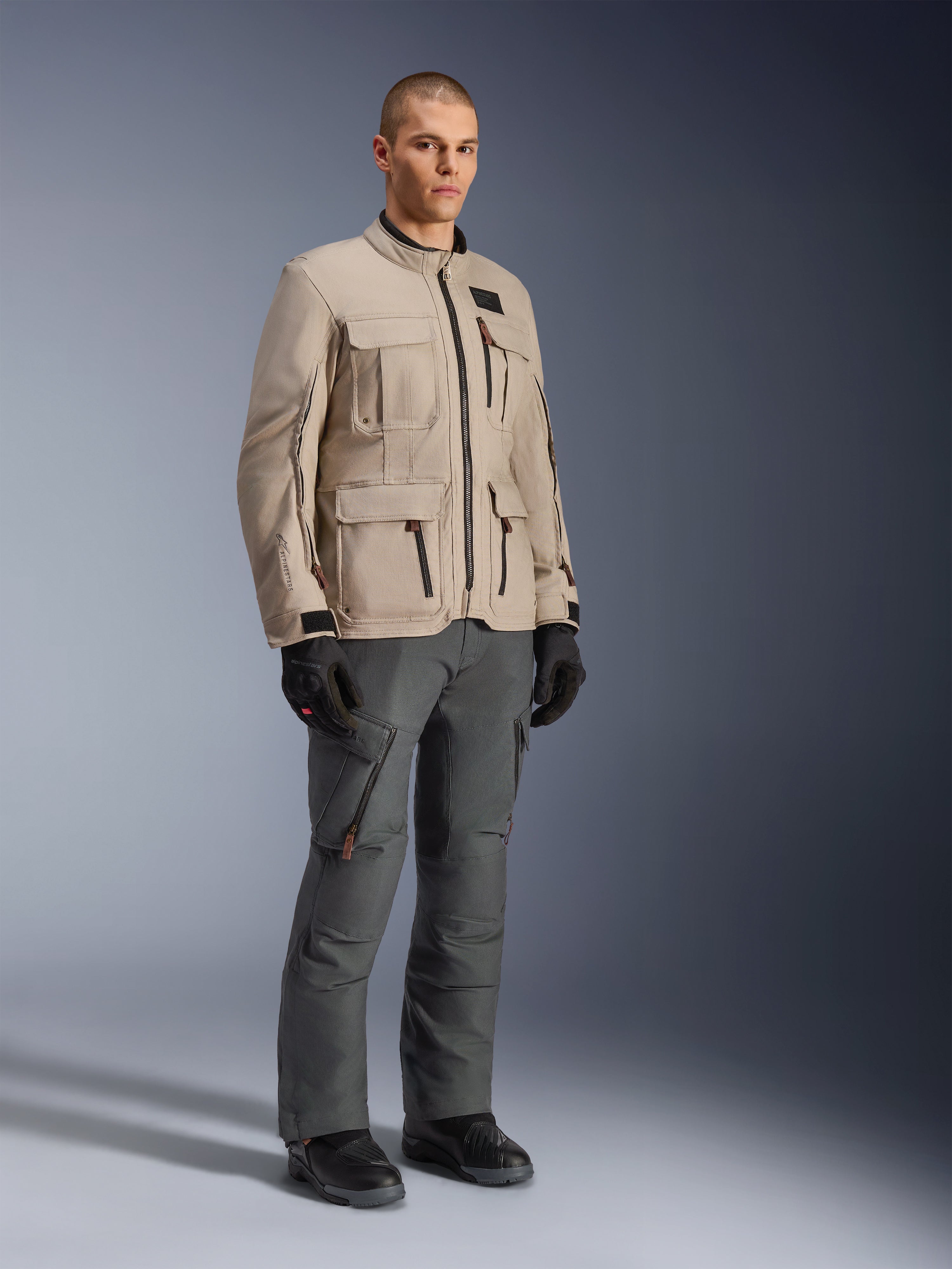 Kurtka Alpinestars Borrego Drystar® w kolorze Warm Gray, przygodowa kurtka motocyklowa o wyglądzie woskowanego płótna z wieloma kieszeniami użytkowymi, noszona przez modela z ciemnoszarymi spodniach motocyklowymi, czarnymi rękawicami i czarnymi butami, portret studyjny całej sylwetki