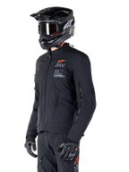 Kurtka Alpinestars AMT-8 Stretch Drystar® XF, czarna, noszona przez motocyklistę z dopasowanymi spodniami adventure i rękawicami, czarny kask motocrossowy i gogle, zapinane na zamek otwory wentylacyjne na klatce piersiowej i rękawach, branding Alpinestars Motion Technology z pomarańczowym logo, wysokiej jakości odzież adventure-touring