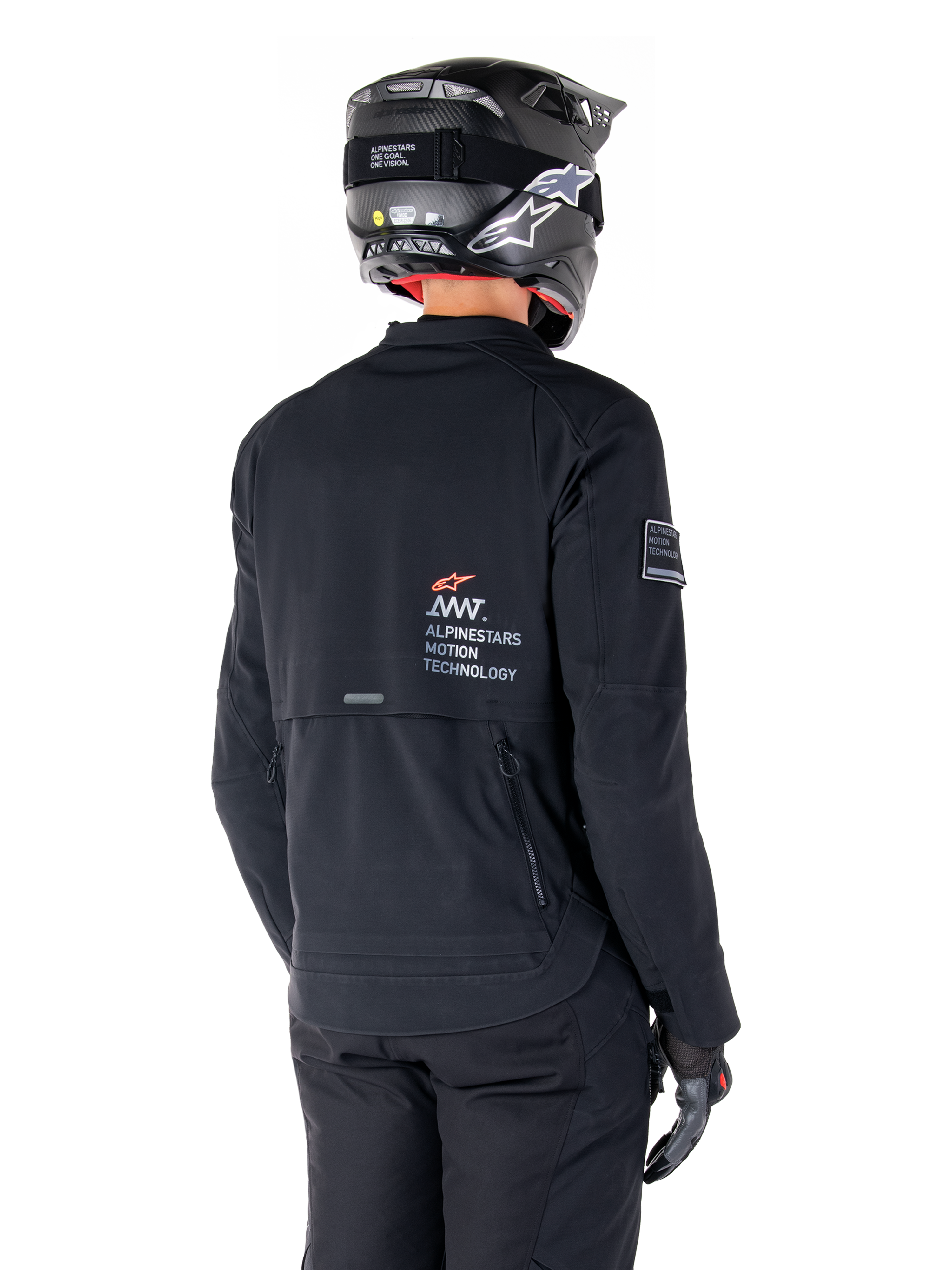 Widok z tyłu motocyklisty w czarnej kurtce Alpinestars AMT-8 Stretch Drystar® XF, z logo „AMT Alpinestars Motion Technology” na plecach, techniczną wentylacją na zamki, w czarnym kasku motocyklowym i rękawicach