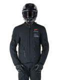Kurtka Alpinestars AMT-8 Stretch Drystar® XF, czarna kurtka motocyklowa, z pomarańczowym logo Alpinestars i szarymi oznaczeniami AMT, wysoki kołnierz z przednim zamkiem i otworami wentylacyjnymi, noszona z czarnym kaskiem i rękawicami, ochronna odzież turystyczna adventure
