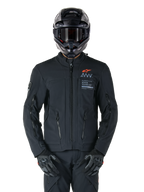 Kurtka Alpinestars AMT-8 Stretch Drystar® XF, czarna kurtka motocyklowa, z pomarańczowym logo Alpinestars i szarymi oznaczeniami AMT, wysoki kołnierz z przednim zamkiem i otworami wentylacyjnymi, noszona z czarnym kaskiem i rękawicami, ochronna odzież turystyczna adventure