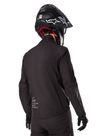 Alpinestars AMT-8 Stretch Drystar® XF Liner, czarna wodoodporna podszewka kurtki, widok z tyłu na motocyklistę w czarnym kasku i rękawicach, z logo Alpinestars Motion Technology na dole pleców, minimalistyczny i funkcjonalny design do jazdy adventure