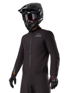Motocyklista w podpince Alpinestars AMT-8 Stretch Drystar® XF Liner, czarna wiatroszczelna i wodoodporna elastyczna kurtka, z logo AMT na klatce piersiowej i oznaczeniem Drystar XF na rękawie, w zestawie z czarnym kaskiem adventure i rękawicami ochronnymi.