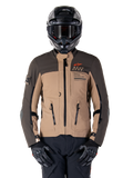 Kurtka Alpinestars AMT-8 Stretch Drystar XF, motocyklowa kurtka turystyczna adventure, ciemne khaki i ciemny brąz, z pomarańczowym logo Astars i czarnymi zamkami, noszona z czarnym kaskiem adventure i rękawicami, pełny zestaw odzieży motocyklowej