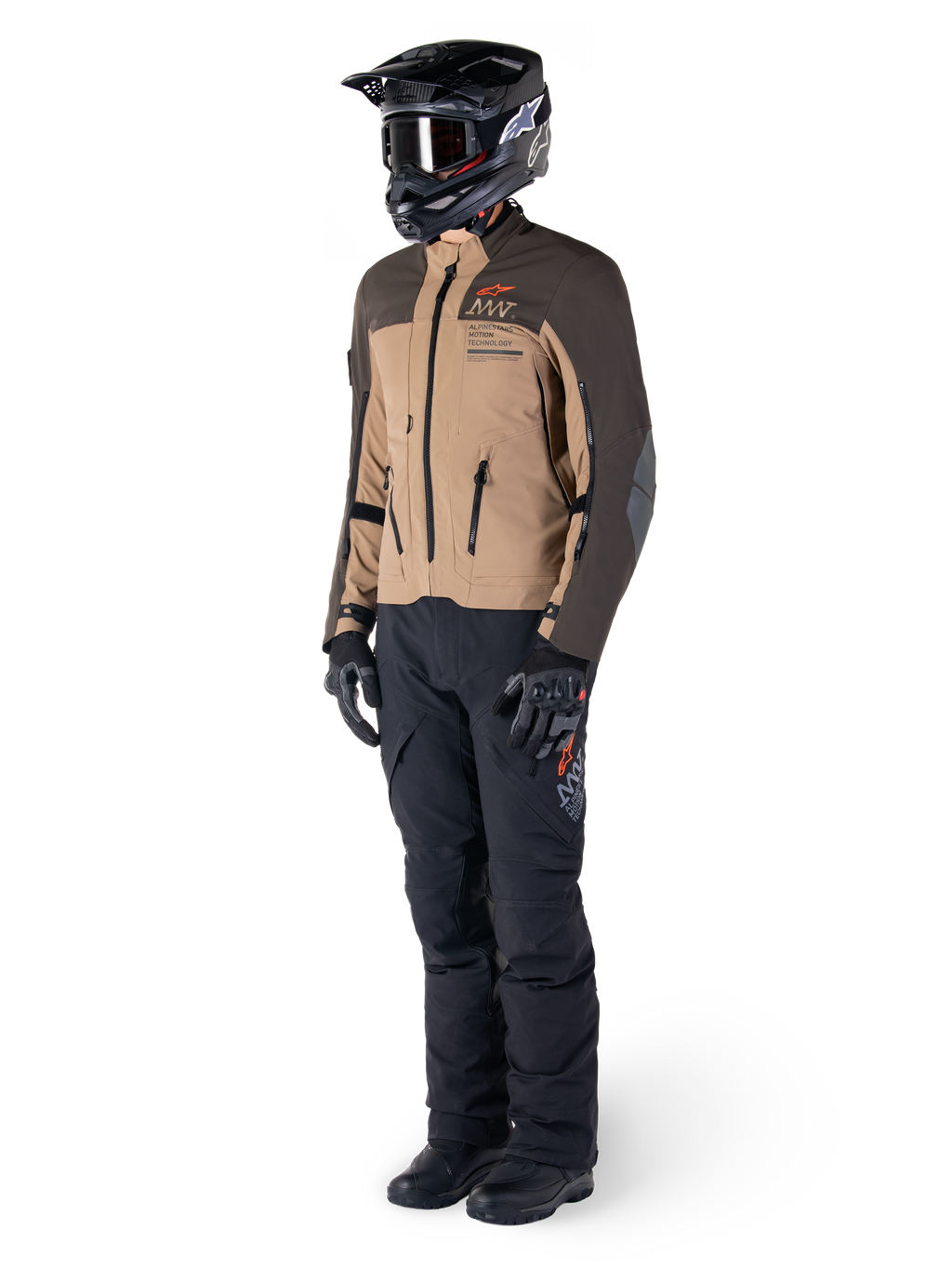 Motocyklista adventure w pełnym rynsztunku, kurtka Alpinestars AMT-8 Stretch Drystar® XF w kolorze ciemnobrązowym i ciemnym khaki, czarne spodnie adventure, czarny kask, rękawice i buty, pomarańczowe logo AMT oraz techniczne zamki wentylacyjne do turystyki długodystansowej