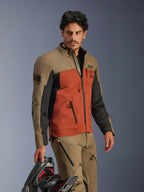 Kurtka Alpinestars AMT-8 Stretch Drystar® XF, Walnut Rust Black, przygodowa kurtka motocyklowa z wysokowydajnego elastycznego materiału, noszona z dopasowanymi orzechowymi spodniami, w ręku czarny kask, odzież adventure touring do jazdy długodystansowej