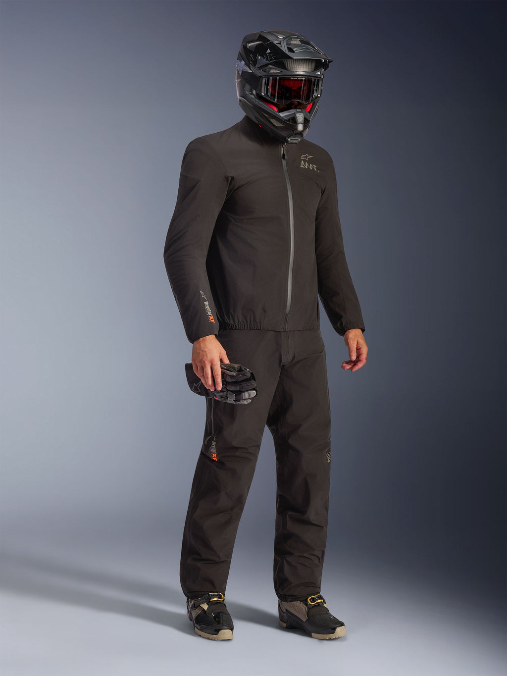 Kurtka Alpinestars AMT-8 Stretch Drystar® XF w kolorze czarnym, noszona jako element pełnego stroju motocyklowego z dopasowanymi spodniami i czarnym kaskiem, elegancki tekstylny design z pomarańczowym logo Drystar XF na rękawie, idealna do turystyki adventure.