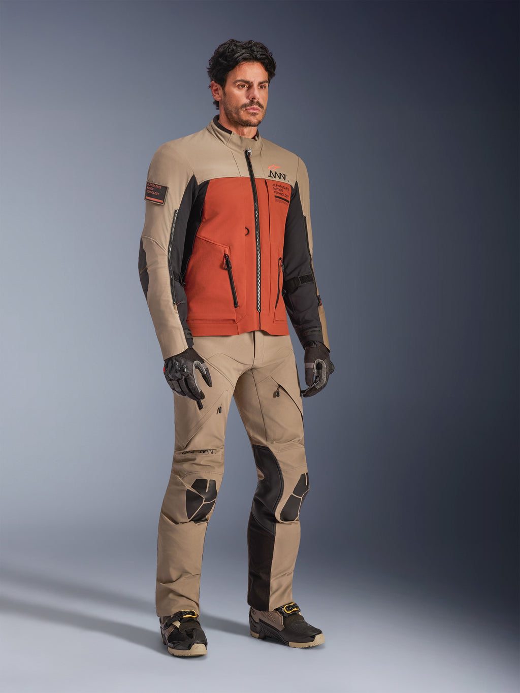 Kurtka Alpinestars AMT-8 Stretch Drystar® XF, kurtka motocyklowa adventure w kolorze Walnut Rust Black, noszona z pasującymi spodniami technicznymi i rękawicami, wielobarwny design z beżowymi, rdzawymi i czarnymi panelami, zaprojektowana do dalekobieżnej turystyki motocyklowej.