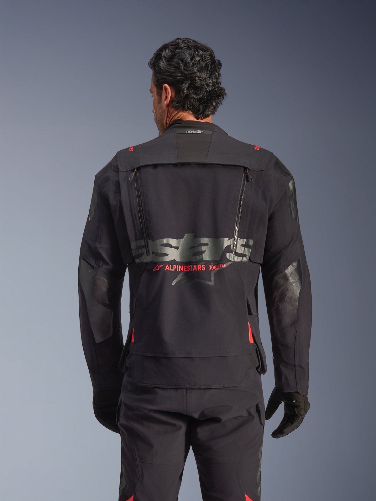 Kurtka Alpinestars Halo Pro Drystar® XF Laminated w kolorze Black Dark Shadow, widok z tyłu na modelu, techniczny panel wentylacyjny na plecach z zamkami, czerwone akcenty logo Alpinestars oraz branding Tech-Air® na karku, modułowa odzież motocyklowa typu adventure.