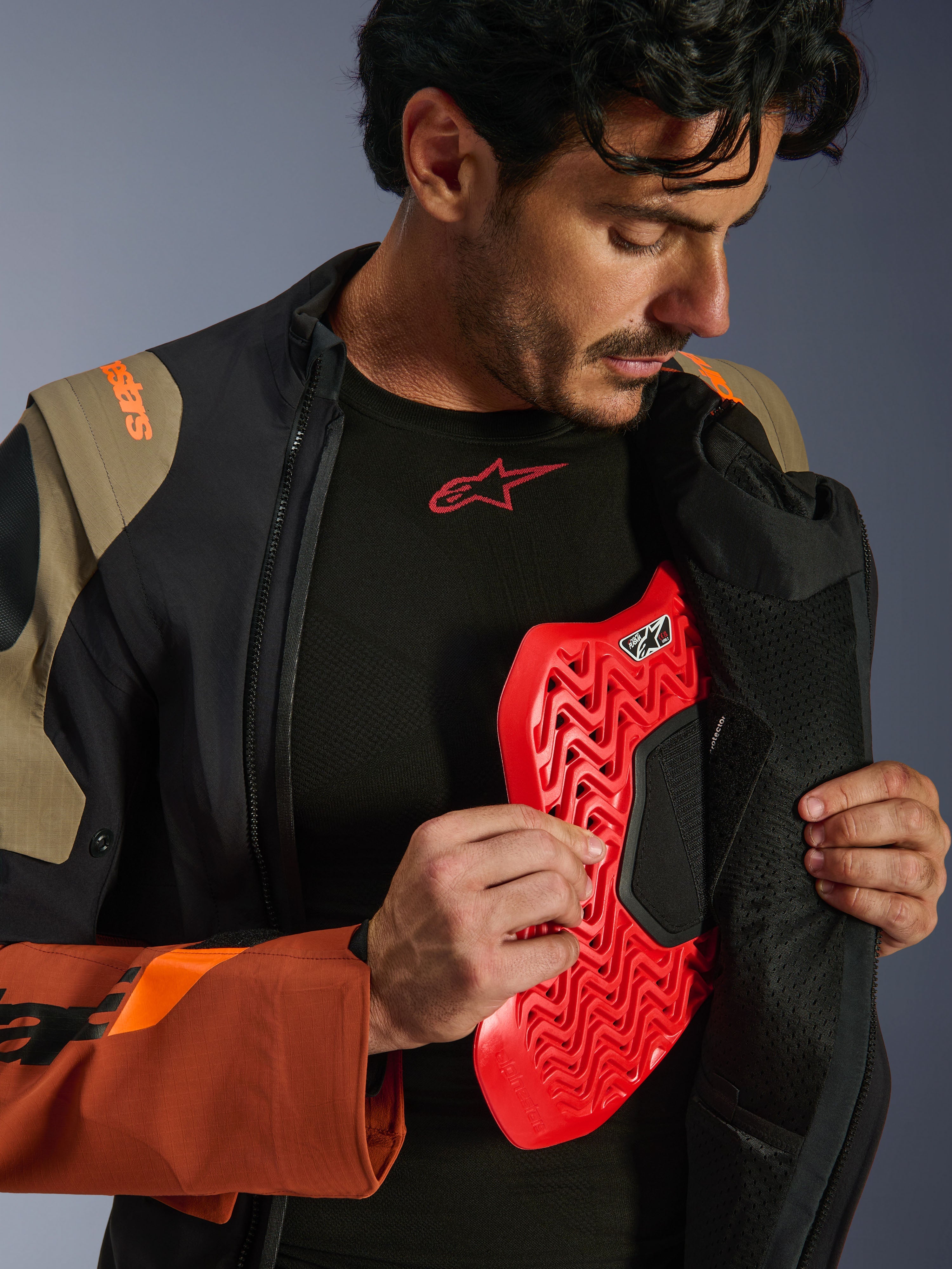 Alpinestars Halo Pro Drystar® XF Laminated Jacket in Walnut Rust Black, lifestyle shot of a male model unzipping the adventure jacket to reveal a red chest protector insert, featuring orange branding on the shoulder and a black base layer with a red logo.