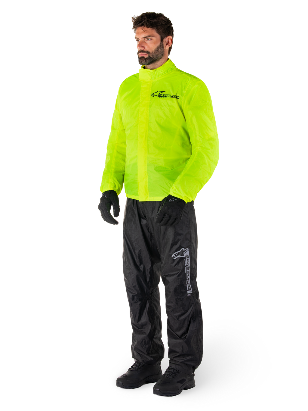 Mężczyzna ubrany w kurtkę Alpinestars Hurricane Rain V2 w kolorze Yellow Fluo i spodnie Hurricane Rain V2 w kolorze czarnym, wodoodporna motocyklowa odzież przeciwdeszczowa o wysokiej widoczności, odblaskowe logo na klatce piersiowej i udzie, kompletny zestaw z czarnymi rękawicami i butami.