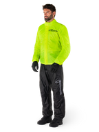 Mężczyzna ubrany w kurtkę Alpinestars Hurricane Rain V2 w kolorze Yellow Fluo i spodnie Hurricane Rain V2 w kolorze czarnym, wodoodporna motocyklowa odzież przeciwdeszczowa o wysokiej widoczności, odblaskowe logo na klatce piersiowej i udzie, kompletny zestaw z czarnymi rękawicami i butami.