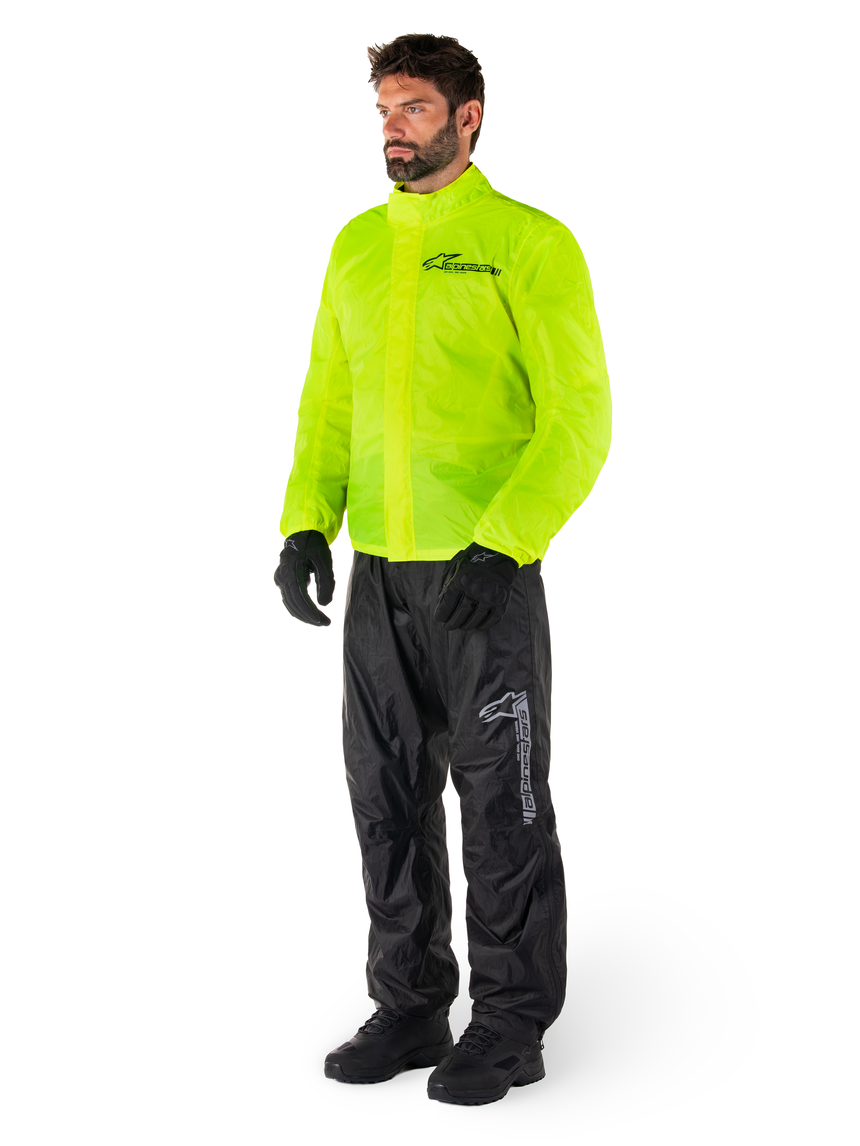 Mężczyzna ubrany w kurtkę Alpinestars Hurricane Rain V2 w kolorze Yellow Fluo i spodnie Hurricane Rain V2 w kolorze czarnym, wodoodporna motocyklowa odzież przeciwdeszczowa o wysokiej widoczności, odblaskowe logo na klatce piersiowej i udzie, kompletny zestaw z czarnymi rękawicami i butami.