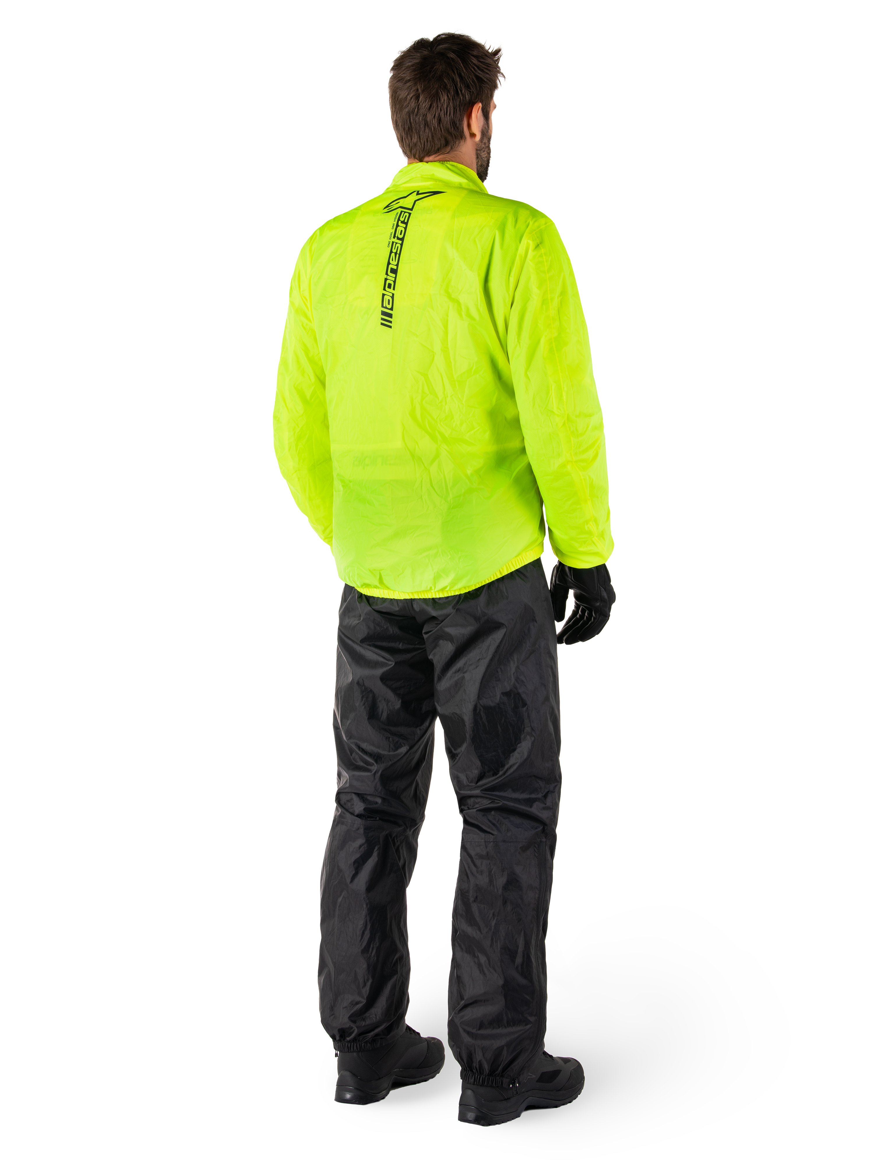 Osoba widoczna od tyłu ubrana w kurtkę Alpinestars Hurricane Rain V2 w kolorze Yellow Fluo i spodnie Hurricane Rain V2 w kolorze czarnym, pionowe logo na plecach kurtki, wodoodporna motocyklowa odzież przeciwdeszczowa, czarne rękawice i buty